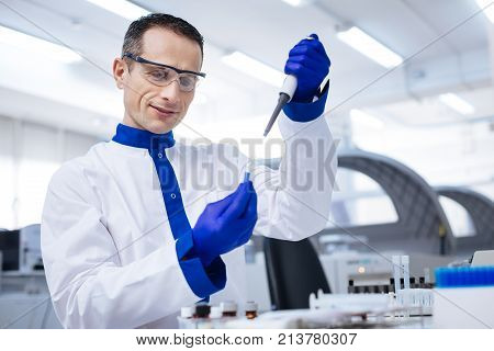 Great sample. Waist up of tranquil enthusiastic male lab assistant  who taking sample from the glassware with blue chemical placed there when standing on the blurred background