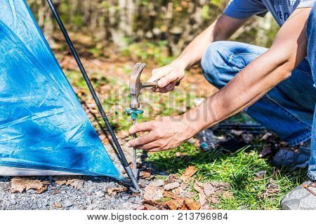 Young Man Setting Up Camp Tent For Camping Trip On Campground By Hammering Nail To The Ground With H