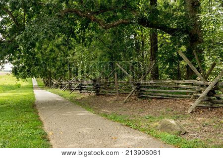 A paved footpath through the countryside beside a split rail fence.