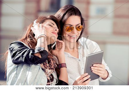 Two beautiful young women walking down the street and listening to music have fun with tablet. Lifestylepeople and technology concept.