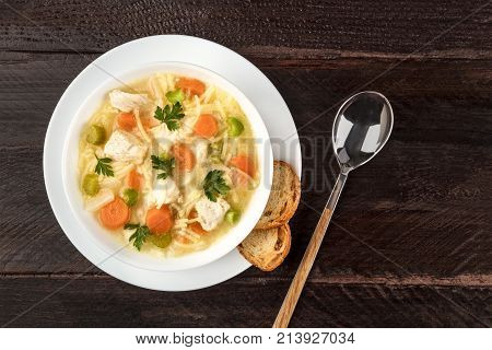 An overhead photo of a plate of chicken, vegetables, and noodles soup, shot from above on a dark rustic texture with slices of bread and a spoon, and a place for text