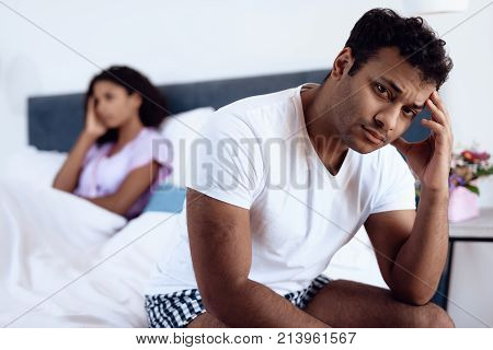 Black man and woman in the bedroom. The man has problems in bed and his woman is not happy with this. She lies behind him in bed, he sits on the edge of the bed with his back to her.