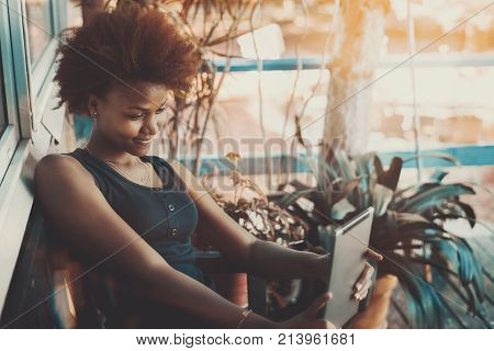 Foxy young African American curly female is sitting on the bench of dacha veranda and taking selfie using front camera of her tablet pc while having online conversating with her friend mate