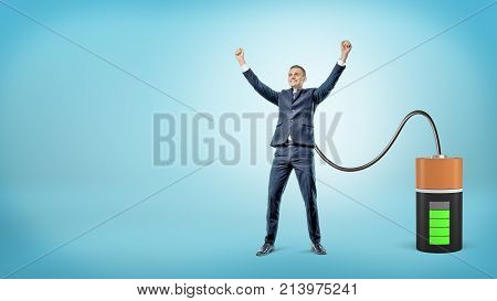 A happy businessman with raised hands is connected to a large battery charging him. Living energy. Power for change. Strength for success.