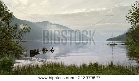 panoramic view of the lake in the summer in the fog. Old broken wooden boat near the shore. Around the highest mountains in the clouds. on the lake floats a small sailboat