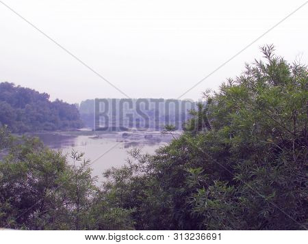 River Landscape With Shores Overgrown With Bushes And Trees With Water, Foggy Horizon And Bright Sky