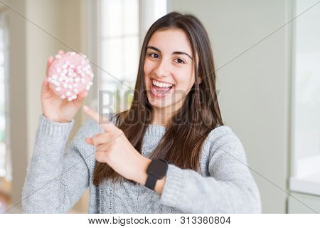 Beautiful young woman eating sugar marshmallow pink donut very happy pointing with hand and finger