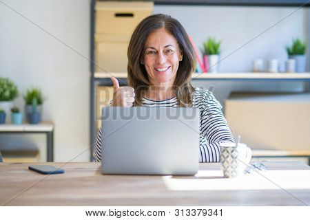 Middle age senior woman sitting at the table at home working using computer laptop doing happy thumbs up gesture with hand. Approving expression looking at the camera showing success.