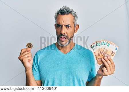 Middle age grey-haired man holding bunch of 50 euro banknotes and bitcoin in shock face, looking skeptical and sarcastic, surprised with open mouth 