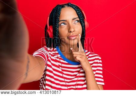 Beautiful hispanic woman taking a selfie photo wearing headphones serious face thinking about question with hand on chin, thoughtful about confusing idea 