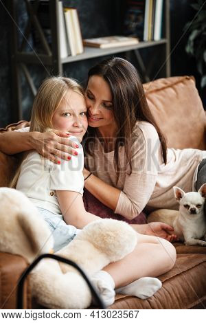Mother and daughter relationship concept. A woman and a teenage girl are sitting in front of the TV on the couch and happy embarcing. The family spends time at home. Little dog lying on laps.