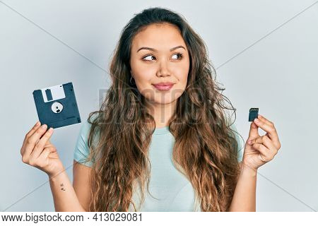 Young hispanic girl holding floppy disk and sdxc card smiling looking to the side and staring away thinking. 