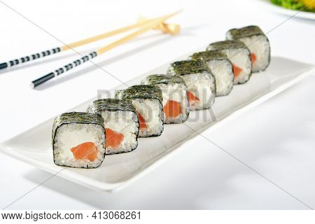 Philadelphia Sushi Roll made with Smoked Salmon. Maki sushi with nori seaweed outside, salmon and cream cheese inside. White plate with wooden chopsticks on white table