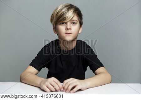 A Serious Boy Of 9 Years Old With A Fashionable Hairstyle In A Black T-shirt Sits At The Table. Dist