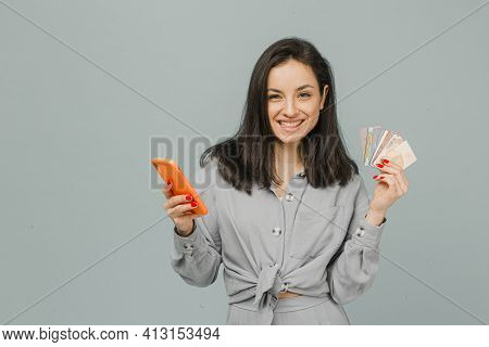 Photo Of Cute Smiling Female Holds Smartphone And Credit Cads. Wears Grey T-shirt, Isolated Grey Col