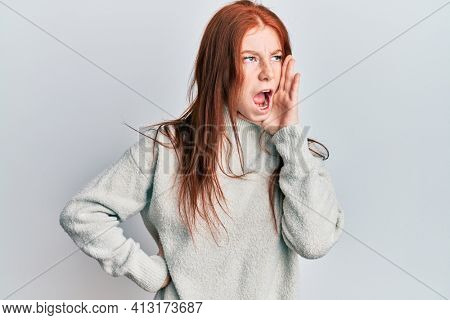 Young red head girl wearing casual winter sweater shouting and screaming loud to side with hand on mouth. communication concept. 