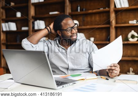 Latin Businessman Sits At Desk In Home Office Near Laptop, Touches Back Of The Head, Thinks Over Str