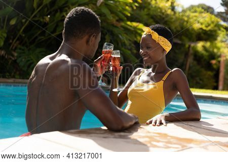African american couple in water drinking drinks by poolside on sunny garden terrace. staying at home in isolation during quarantine lockdown.