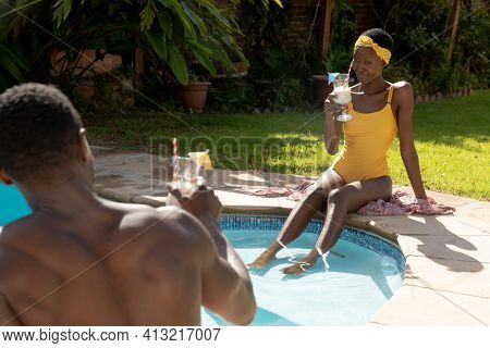 African american couple sitting at the poolside and drinking drinks on sunny garden terrace. staying at home in isolation during quarantine lockdown.
