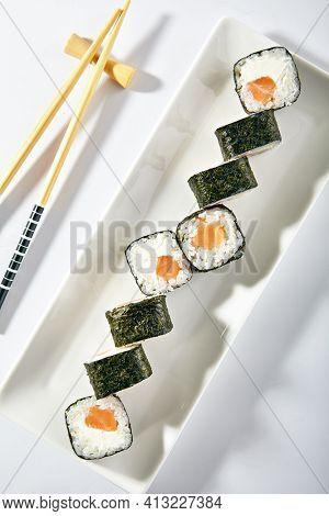 Philadelphia Sushi Roll made with Smoked Salmon. Maki sushi with nori seaweed outside, salmon and cream cheese inside. White plate with wooden chopsticks on white table. Top view