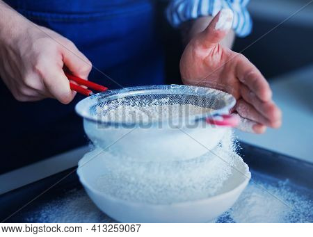 A Man In A Blue Apron Sifts Flour Through A Sieve Into A White Bowl, In Which He Is Going To Mix The