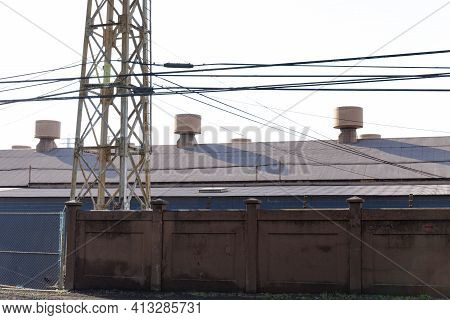 Base Of A Metal Power Line Tower Between A Tall Wall And An Industrial Building With Air Turbines On
