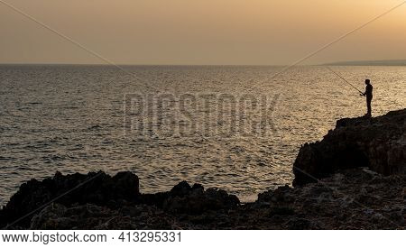 Silhouette Of A Fisher Fisheman Fishing In The Ocean At Sunset. Ayia Napa, Cyprus