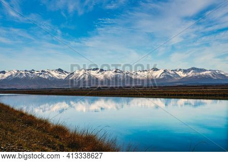 Snow Covered Southern Alps Mountain Range Near Twizel Reflected In The Still Water In The Canal Flow