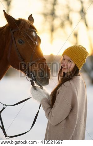 Winter Side Portrait Of Young Smiling Woman And Brown Horse. Woman With Long Hear In Yellow Cap Hold
