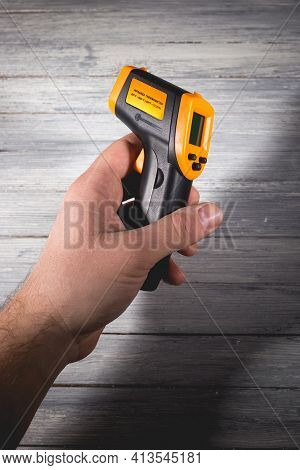 A Man Holds A Yellow-black Pyrometer On A Wooden Background. A Device For Non-contact Temperature Me
