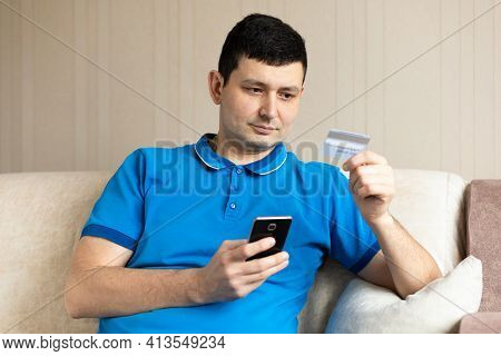 Millennial Man Holding Credit Bank Card And Using Smartphone, Mobile At Home. Online Payment, Purcha