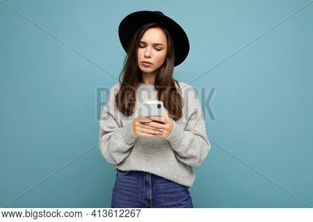 Beautiful Young Brunette Woman Thinking Wearing Black Hat And Grey Sweater Holding Smartphone Lookin