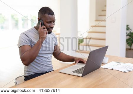 African american man working from home sitting at table using laptop and talking on smartphone,. staying at home in isolation during quarantine lockdown.