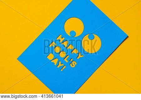 the text happy fools day and a pair of funny eyes cut out in a blue paperboard, against an orange background