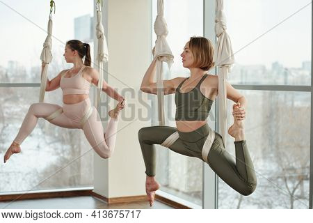 Two young active females in tracksuits sitting on fly yoga hammocks and hanging over floor of gym with their legs bent in knees