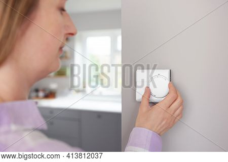 Woman At Home In Kitchen Turning Down Central Heating Thermostat To Save Energy And Money