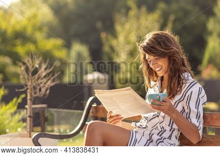 Beautiful Young Woman Spending Sunny Summer Morning Outdoors, Having Breakfast, Reading Newspaper An