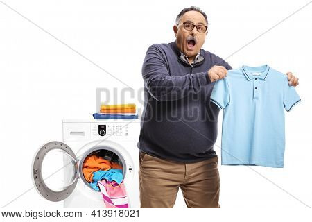 Surprised mature man holding a shrunken t-shirt in front of a washing macine isolated on white back