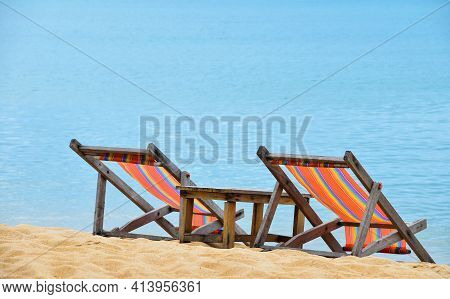 Two Empty Colorful Canvas Lounge Sling Chairs And Wooden Table Over Blue Sea, On Sand Beach Of Resor