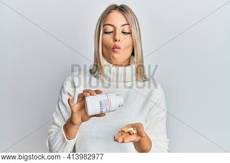 Beautiful blonde woman holding pills making fish face with mouth and squinting eyes, crazy and comical. 
