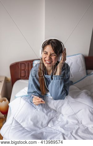 Beautiful Young Woman Sitting In Bed, Relaxing At Home In The Morning, Wearing Headset And Laughing 