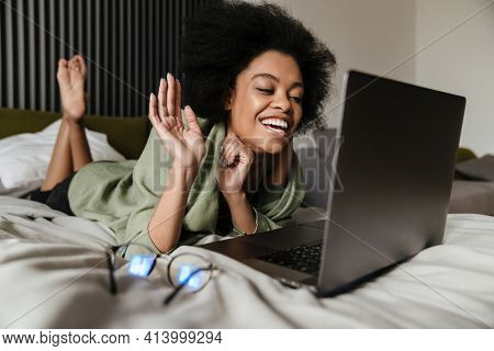 Happy Mid aged african woman using laptop computer while laying in bed at home, video cahtting while self isolating at home, waving hand
