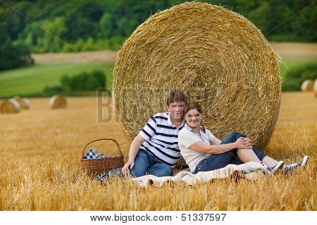 Young Couple Picknicking On Yellow Hay Field On Summer Evening.