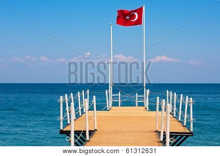 Small wooden pier and red national flag waving on the wind in Kemer - tourist resort on Mediterranean sea in Turkey.