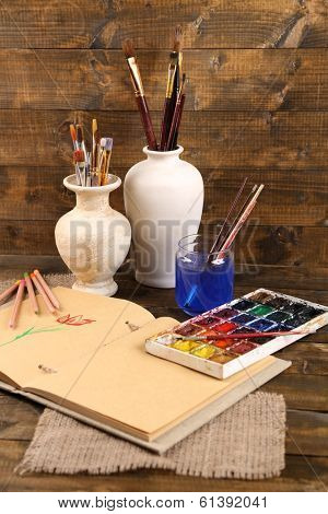 Composition with colorful watercolors, brushes in vase and sketcher on wooden background 