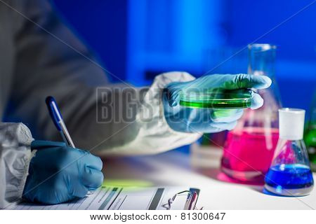 science, chemistry, medicine and people concept - close up of young scientist with chemical sample taking notes on clipboard and making test or research in laboratory