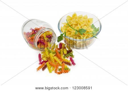 Uncooked dried spiral pasta colored by beetroot spinach paprika carrot grape seeds in jar and next to it bow-tie pasta with a sprig of parsley in glass bowl on a light background