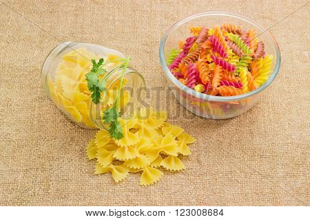 Uncooked dried spiral pasta colored by beetroot spinach paprika carrot grape seeds in glass bowl farfalle pasta with a sprig of parsley in jar and next to it on a sackcloth