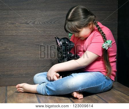 Girl with a black dog on the floor near the wooden walls. The dog - french bulldog puppy. Girl in jeans, barefoot. Dog and little mistress. Girl full