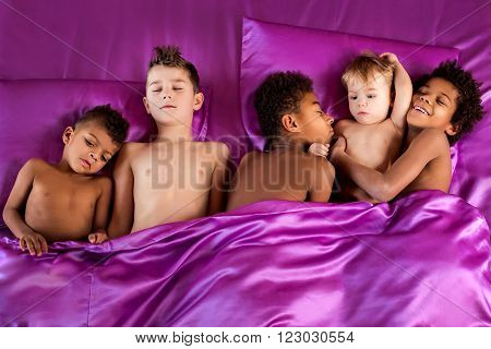 Darkskinned and european boys sleeping. Five children on purple bed. Closely and comfortably. Time to relax.